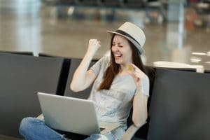jeune-touriste-voyageur-riant-femme-au-chapeau-assis-travaillant-ordinateur-portable-tenant-bitcoin-faisant-geste-du-gagnant-attente-dans-hall-aeroport